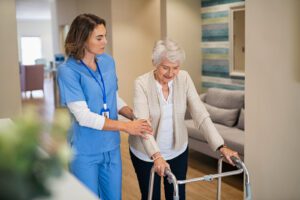 Lovely nurse helping old woman to walk at nursing home with walker. Young nurse helping senior patient using a walking frame to walk in hospital corridor. Caregiver and disabled lady in care facility.